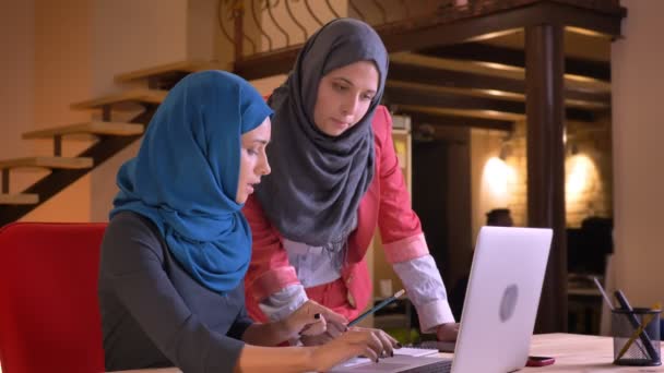 Крупный план портрета молодых красивых мусульманских женщин в хиджабах, обсуждающих командный проект, используя ноутбук в помещении на рабочем месте — стоковое видео