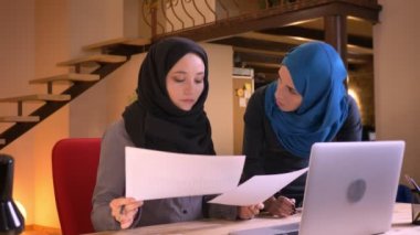 Dizüstü bilgisayar kapalı ofiste kullanarak farklı grafikler üzerinde veri karşılaştırma hijabs içinde iki başarılı Müslüman iş kadınları closeup portresi