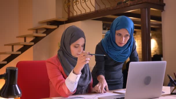Крупный план портрета двух молодых арабских бизнесвумен в хиджабах, спорящих за информацию, представленную на графике, используя ноутбук в помещении на рабочем месте — стоковое видео