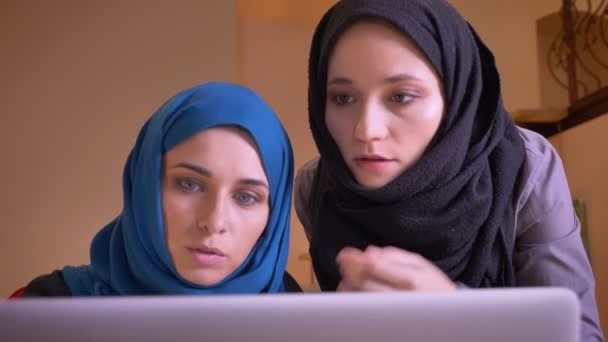 Retrato de cerca de dos jóvenes empresarias musulmanas en hiyabs hablando de la información en el portátil. Jefe instruyendo a una empleada . — Vídeos de Stock