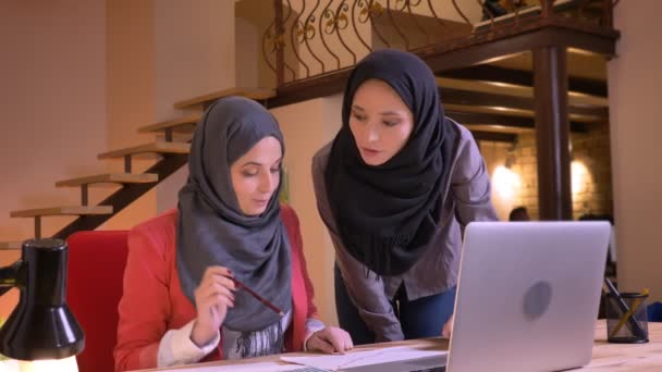 Retrato de close-up de duas jovens empresárias muçulmanas alegres em hijabs discutindo emocionalmente um tópico de trabalho usando o laptop, em seguida, olhando para a câmera e sorrindo — Vídeo de Stock