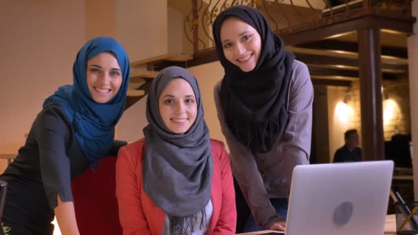 Портрет трех молодых мусульманских офисных работниц в хиджабах, смотрящих прямо в камеру и улыбающихся, находясь перед ноутбуком на рабочем месте — стоковое видео