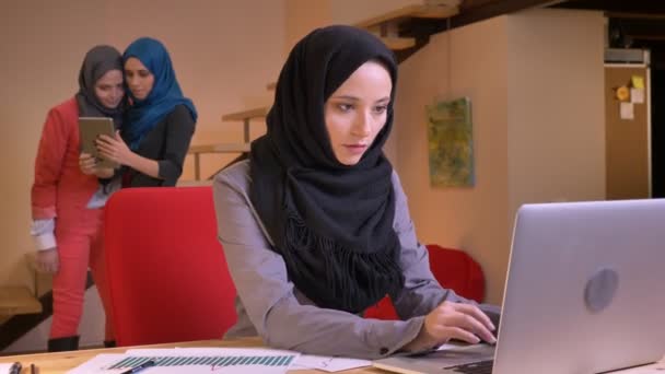Retrato de close-up da jovem trabalhadora de escritório muçulmana concentrada e digitando no laptop com colegas de trabalho dela no fundo tirando selfies no tablet — Vídeo de Stock