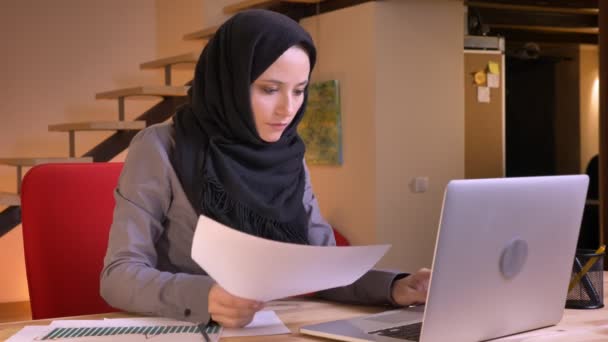 Retrato de cerca de la joven mujer de negocios musulmana hermosa comparando los datos en el gráfico y en la computadora portátil en el lugar de trabajo en interiores — Vídeos de Stock