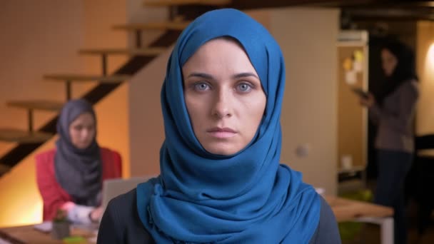 Крупним планом портрет красивої мусульманки в блакитному хіджабі, дивлячись прямо на камеру, що знаходиться на робочому місці в приміщенні — стокове відео
