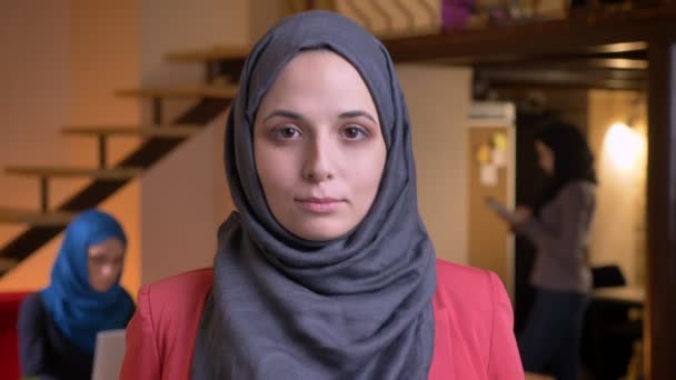 Retrato de cerca de la joven mujer de negocios musulmana hermosa en hijab gris mirando directamente a la cámara que está en el lugar de trabajo en el interior — Vídeo de stock