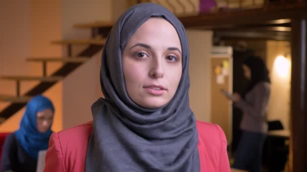 Closeup portrait of young beautiful muslim woman in gray hijab looking thoughtfully in contemplation at camera being on the workplace indoors — Stock Video