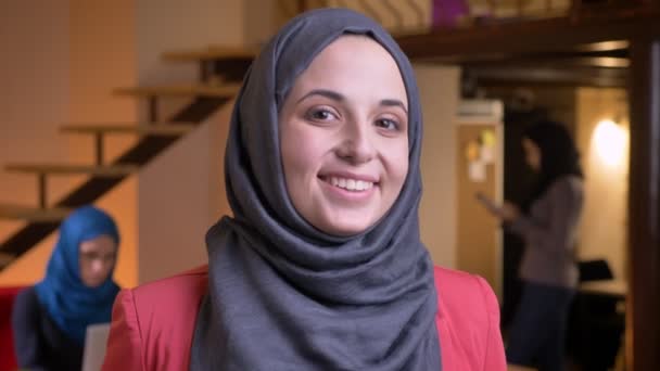 Retrato de close-up de jovem mulher de negócios muçulmana positiva em hijab cinza olhando diretamente para a câmera e sorrindo alegremente estar no local de trabalho — Vídeo de Stock