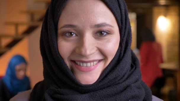 穿着黑色头巾的年轻有趣的穆斯林上班族的特写镜头肖像看着相机, 在室内的工作场所快乐地笑着 — 图库视频影像