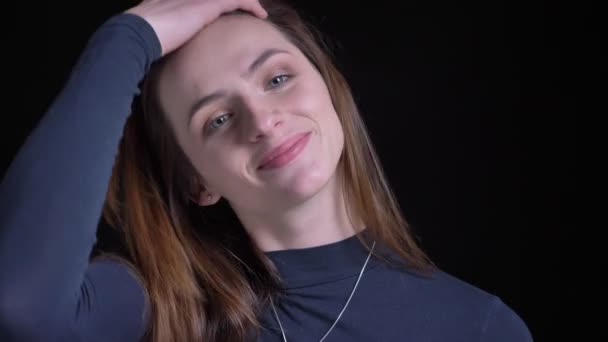 Close-up portrait of young and slim brunette caucasian girl fixing her hair and watching smilingly into camera on black background. — Stock Video