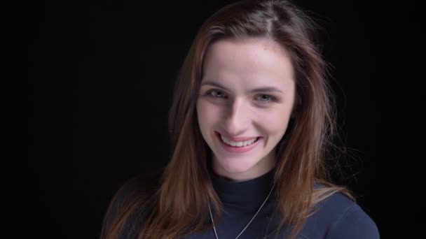 Close-up portret van jonge en slanke brunette Kaukasische meisje glimlachend bescheiden in camera op zwarte achtergrond. — Stockvideo
