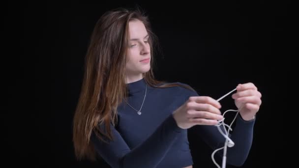 Portrait of young and slim brunette caucasian girl unscrambling earphones on black background. — Stock Video