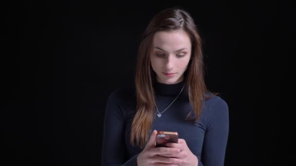 Portrait of young and slim brunette caucasian girl attentively watching into smartphone on black background. — Stock Video
