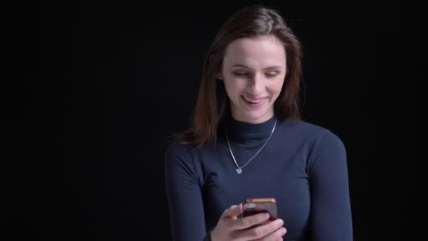 Porträt eines jungen und schlanken brünetten kaukasischen Mädchens, das ins Smartphone schaut und in die Kamera auf schwarzem Hintergrund lacht. — Stockvideo