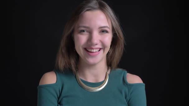 Nahaufnahme Porträt eines jungen brünetten kaukasischen Mädchens, das nachdenklich nickt und auf schwarzem Hintergrund in die Kamera lächelt. — Stockvideo