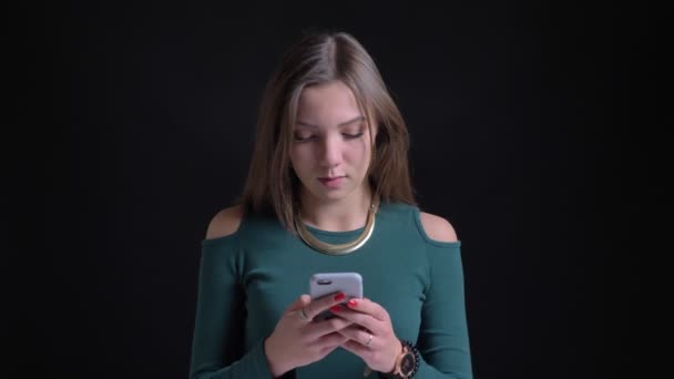 Portrét mladé brunetka kavkazské sledování do smartphone radostně na černém pozadí. — Stock video