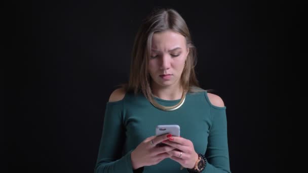 Portret van jonge brunette Kaukasische meisje lachen kijken in smartphone op zwarte achtergrond. — Stockvideo