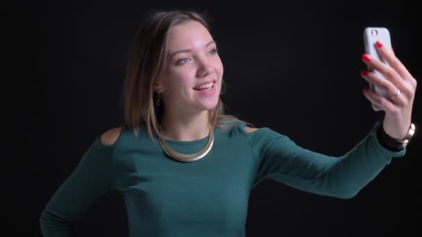 Portrait of young brunette caucasian girl talking in videochat using smartphone joyfully on black background. — Stock Video