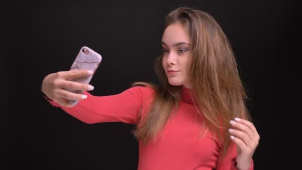 Closeup portrait of young beautiful caucasian female taking selfies on the phone tossing her hair and posing in front of the camera — Stock Video