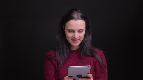 Närbild porträtt av unga kaukasiska kvinna med svart hårfärg använder tabletten och skrattar med bakgrund som isolerade på svart — Stockvideo
