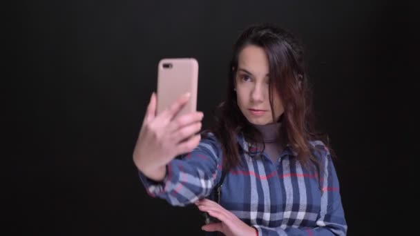 Primer plano retrato de joven caucásica hembra tomando selfies en el teléfono con el fondo aislado en negro — Vídeo de stock