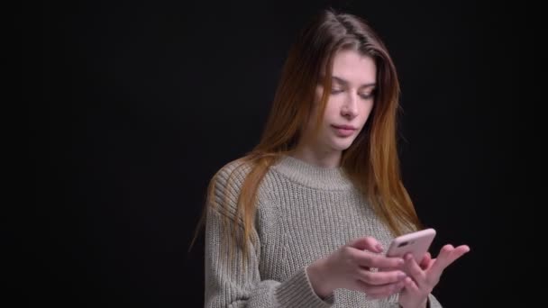 Close-up retrato de jovem muito caucasiano feminino casualmente mensagens de texto no telefone com fundo isolado no preto — Vídeo de Stock