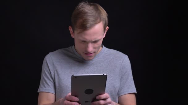 Closeup portret van knappe Kaukasische jongeman te typen op de tablet voor de camera met geïsoleerd op zwarte achtergrond — Stockvideo