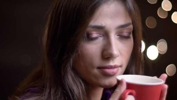 Primo piano ritratto di giovane bella femmina caucasica con una tazza di tè caldo rivolgendosi alla fotocamera sorridente e pensieroso in avanti — Video Stock