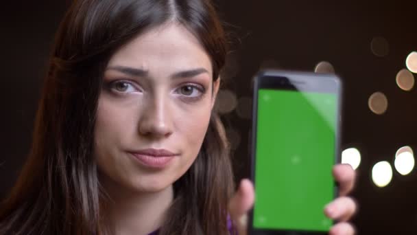 Primo piano ritratto della giovane bella ragazza caucasica che tiene un telefono e mostra lo schermo verde alla fotocamera gesticolando un pollice verso l'alto — Video Stock