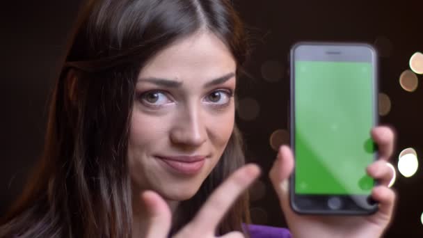 Retrato de close-up de jovem mulher caucasiana encantadora segurando um telefone apontando na tela verde sorrindo e mostrando o polegar para cima — Vídeo de Stock