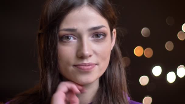 Detailní portrét mladé docela kavkazský dívky s úsměvem a stanovení její vlasy při pohledu kamery s noční světla na pozadí — Stock video