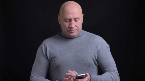 Крупный план портрет взрослого белого лысого мужчины, использующего телефон, а затем смотрящего прямо в камеру на фоне изолированного на черном фоне — стоковое видео