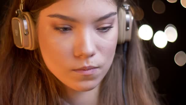 Detailní portrét vážné mladé krásné kavkazský dívky ve sluchátkách poslouchat hudbu na rozmazané světla pozadí. — Stock video
