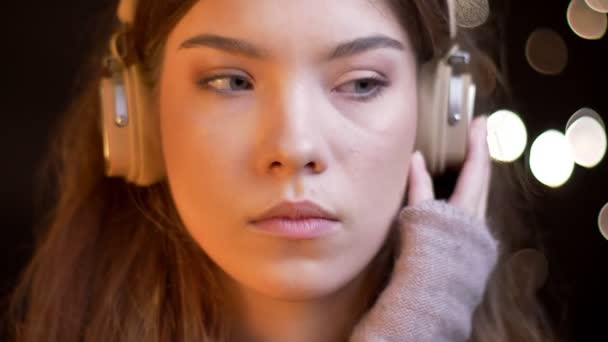 Close-up πορτρέτο του όμορφη Καυκάσιος κοπέλα με ακουστικά που άκουγε προσεκτικά τη μουσική στη θολή φώτα φόντο. — Αρχείο Βίντεο