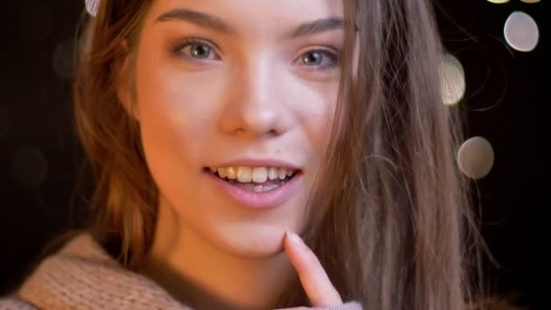 Close-up portret van de jonge Kaukasische meisje glimlachend in de camera kijken en denken over iets op wazig lights achtergrond. — Stockvideo