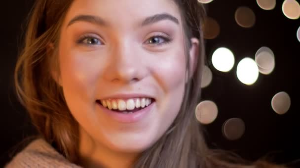 Крупним планом портрет молодої кавказької дівчини сором'язливо сміється в камеру на розмитому тлі вогнів . — стокове відео