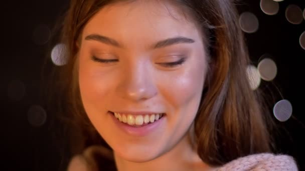 Close-up ritratto di carino giovane ragazza caucasica sorridente umilmente in macchina fotografica su sfondo luci sfocate . — Video Stock