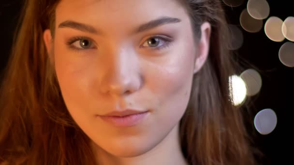 Close-up ritratto di giovane ragazza caucasica guardando modestamente in macchina fotografica su sfondo luci sfocate . — Video Stock