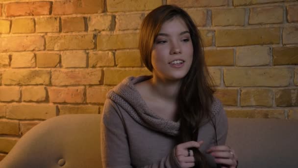 Young and beautiful caucasian girl sitting on sofa and watching thoughtfully into camera on bricken wall background. — Stock Video