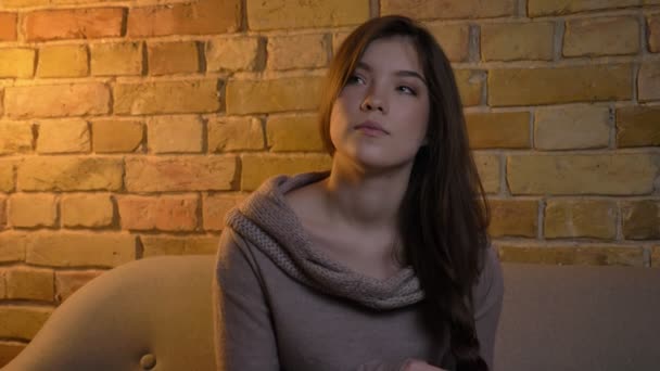 Young and beautiful caucasian girl making a pigtail and watching calmly into camera on bricken wall background. — Stock Video