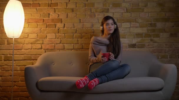 Young and beautiful caucasian girl in headphones sitting on sofa with red cup and dancing on bricken wall background. — Stock Video