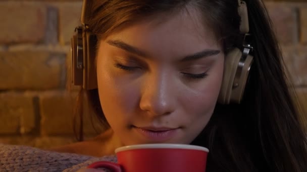 Close-up portret van de jonge Kaukasische meisje in hoofdtelefoons met rode kop, genieten van de drank op bricken muur achtergrond. — Stockvideo