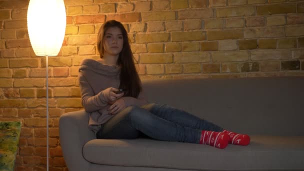 Portret, młoda i piękna, kaukaski dziewczyny, siedząc na kanapie i oglądania telewizji uważnie na przytulnym domu tło. — Wideo stockowe