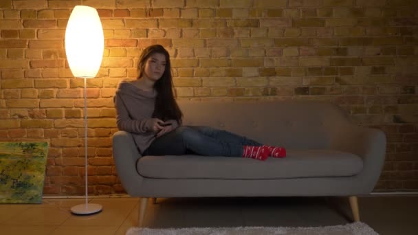 Portrait of young and beautiful caucasian girl sitting on sofa and switching the channels on TV on cosy home background. — Stock Video