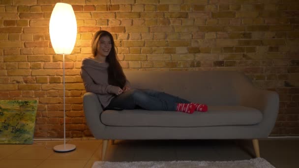 Портрет молодой и красивой кавказской девушки, сидящей на диване и смотрящей телевизор и смеющейся на уютном домашнем фоне . — стоковое видео