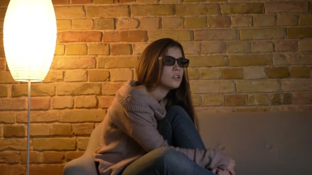 Portrait de jeune fille caucasienne effrayée en lunettes 3D assise sur le canapé et regardant le film d'horreur à la télévision dans un cadre confortable à la maison . — Video
