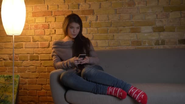 Portrait of young caucasian girl sitting on sofa and watching into the smartphone attentively on cosy home background. — Stock Video