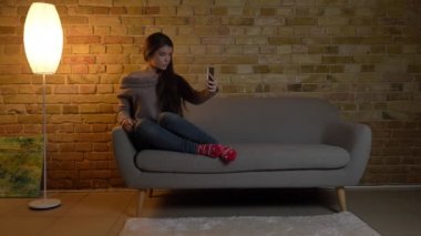 Selfie-fotoğrafları Smartphone'da yapma ve rahat ev arka plan üzerinde gülümseyen kanepede oturan genç beyaz kız portresi.
