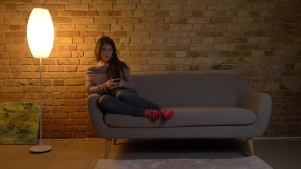 Portret młodej dziewczyny kaukaski, siedząc na kanapie i oglądać do smartfona uśmiechem na przytulnym domu tło. — Wideo stockowe