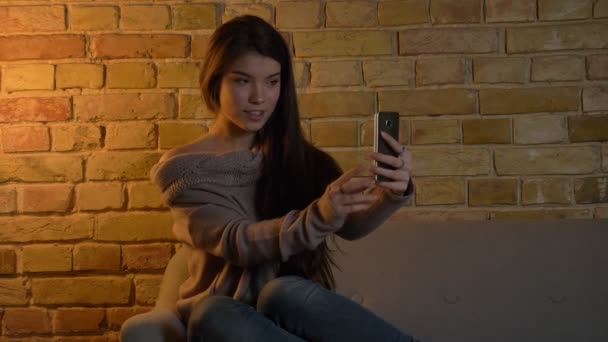 年轻的高加索女孩坐在沙发上, 在舒适的家庭背景下在智能手机上制作自拍照片的肖像. — 图库视频影像